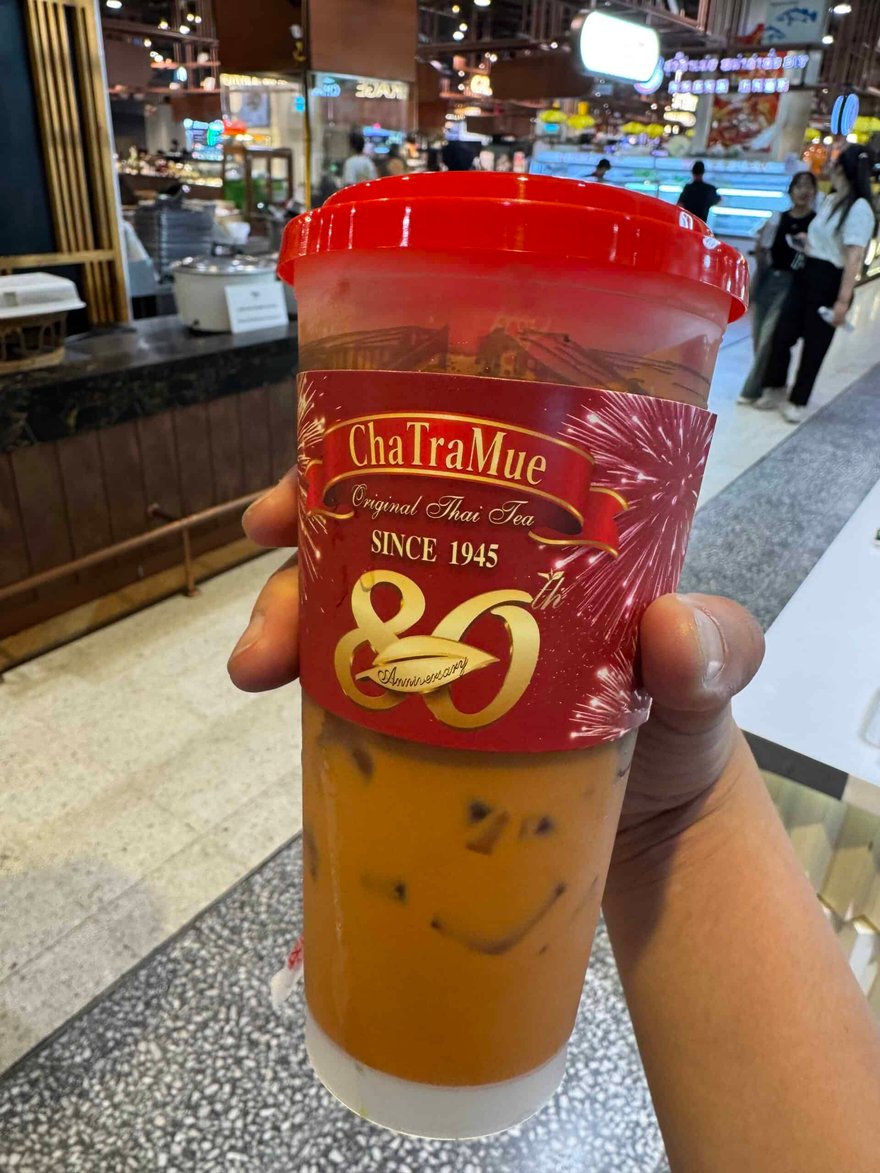 一隻手拿著 ChaTraMue 80 週年紀念杯，紅色杯蓋與杯身，下方是橘色泰式奶茶