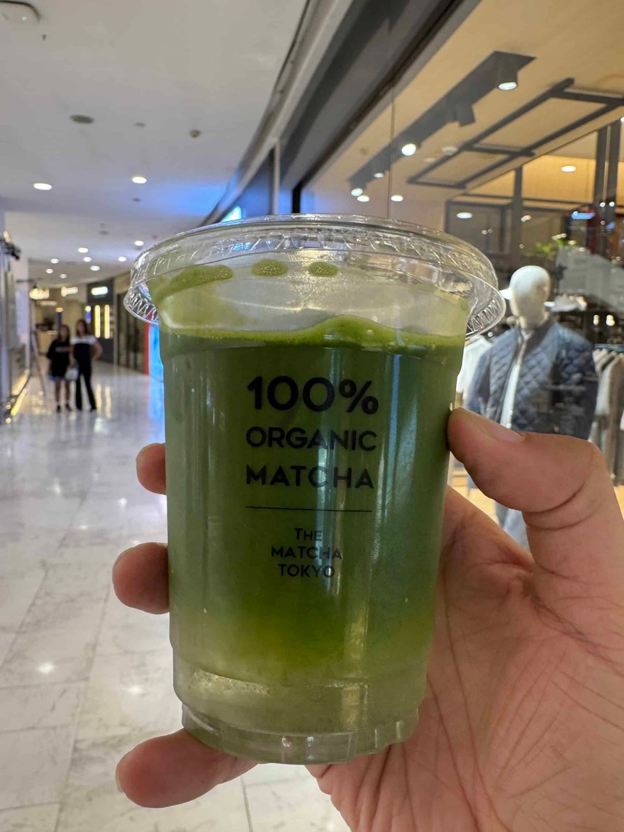 一隻手拿著 The Matcha Tokyo 的綠色抹茶飲料，杯身印有 100% ORGANIC MATCHA，背景是購物中心走道
